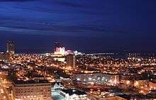 An illuminated cityscape alongside water seen at night from the air.