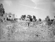 Soldiers advance across rocky ground towards a stone wall and gate