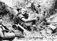 Black and white photo of six men wearing military uniform. The man in the centre of the photograph is sitting on the ground wearing a radio headset, and four of the other men are sitting or crouching