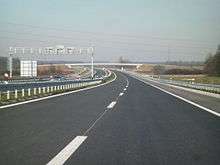 A view of four lane motorway with a central reservation, taken from a carriageway level. Back side of a gantry carrying variable traffic signs is visible on the left hand side carriageway.