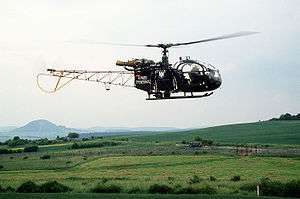 Green-painted helicopter with "Bundesgrenzschutz" on the side flying parallel to a border fence with a gate in it, behind which are two East German soldiers and a canvas-sided truck.