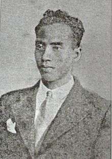 A faded image of a man with short, curly hair in a suit and tie.