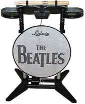 A drum set video game controller with four elevated drum pads mounted on a frame, along with a bass pedal attached to a bottom crossbar. A thin panel, stating "Ludwig" and "The Beatles" is mounted to the front. The drum pads are colored in a metallic gray pattern, while most of the rest is either black or gray.