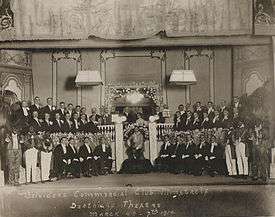 Group photo on a theatre stage of formally dressed men standing in three rows, one woman with small group at centre. The blackfaced end men are costumed.