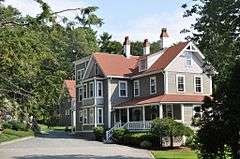 A rambling gray and white colonial house with a porch sheltering the main entrance on the side of the house.