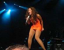 A brunette woman with dark skin wears an orange dress. She holds a microphone and she performs a song on a stage.