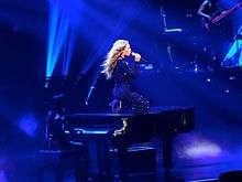 A woman kneels upon a piano whilst singing