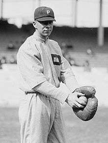 A black-and-white image of a man wearing a white old-style baseball uniform and holding a baseball with his right hand in the catcher's mitt on his left hand