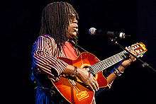 A man with hair braids is wearing sunglasses and playing a guitar.