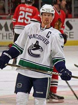 An ice hockey player wearing a white and blue jersey with a logo of a stylized orca in the shape of a "C".  He stands relaxed on the ice looking forwards.