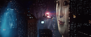 Screenshot of a police spinner flying through a cityscape next to a large building which has a huge face projected onto it. In the distance a screen can be seen with writing and pictures on it