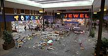 Film screenshot showing a police car driving through a shopping mall: Scattered items are present on the floor and people are running away from the vehicle. Stores visible in the mall include Toys 'R' Us and Jewel.