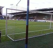 In the foreground is a football net. Through the net, a football stand with wooden seats can be seen in addition to part of a grass football pitch. In the top left corner there is a floodlight pylon.