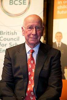 A photograph of a bald man looking at the camera. The man is wearing a black suit, white shirt, and a red tie.