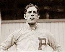 A black-and-white photograph of a dark-haired man in a white baseball jersey standing with arms akimbo