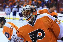 An ice hockey goal tender is stood in front of a goal, peering to his left. He is wearing a black uniform with orange and white piping, and a logo on his chest.