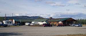 A low-slung wooden structure is surrounded by vehicles on a dirt parking lot