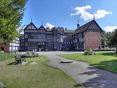 Photo a long, two-storied house with exposed wooden beams, many gables and small-paned windows. In the foreground is a lawn and trees are on the left.