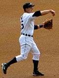 Baseball player in white uniform in a baseball throw followthrough position.  His arm is forward with his hand down and his left hand is gloved. His left foot is planted and his right foot is in the air behind him.