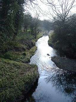 A steam flowing away for the camera, wooded banks on either side, bright sky in the upper portion of the picture