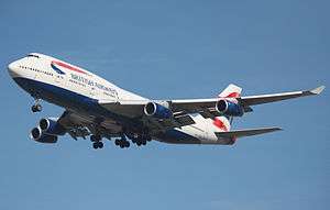 A large white and blue four-engine jet airliner with union tail fin, with landing gear extended