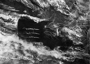 Black and white photo of a four-engined aircraft viewed from above while flying over a narrow body of water