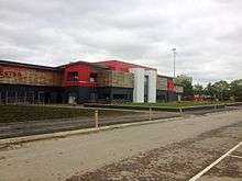 Broadhurst Park football ground on an overcast day, with an access road in the foreground. Visible are two white towers forming the front of the stadium, with a balcony to the left of the left tower. One of the unlit stadium floodlights is visible in the background.