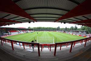 A view of a football pitch from behind the goals. There is a stand with red, white and black seating to the left. There are covered stands without terracing on the right and beyond the pitch. In the forefront is terracing with red barriers and a roof above. Green trees can be seen beyond the stadium.