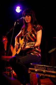 A woman with long black hair wearing a red and black striped singlet. She is seated and playing a guitar in front of a microphone.