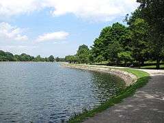 The curving shoreline of a body of water is lined with roughly dressed granite stones.  A walkway follows the curve of the shore, with a trees providing shade.