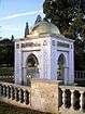 Roughly cubic white stone structure, topped by a gilded dome. Each of the four faces of the structure features a large pointed archway, and the two arches facing the camera also feature ornate Arabic calligraphy.