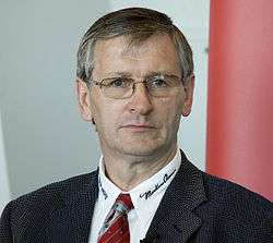 A portrait photograph of a grey-haired man wearing glasses and a black suit.