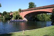 A wide tree-lined river on the left has a green bank in the right-foreground and is crossed by a low two brick-arched bridge. A tree in the middle of the river obscures part of the bridge.