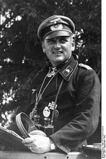 A man wearing a peaked cap, black military uniform with various military decorations.