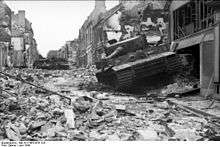 Three stationary tanks, one in the foreground and two in the rear, in a heavily damaged street; rubble covers the road.