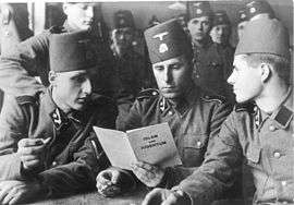 three soldiers in SS uniform and wearing fez headgear reading a pamphlet