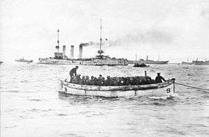 A small white boat filled with soldiers passes in front of a large warship with three tall funnels and several smaller transport ships.