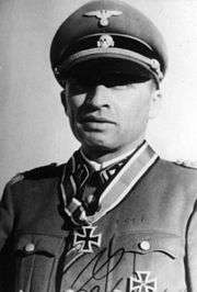 A man wearing a military uniform, peaked cap and a neck order in the shape of a cross. His cap has an emblem in shape of a human skull and crossed bones.