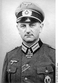 The head and shoulders of a man. He wears a peaked cap and a military uniform and an Iron Cross displayed at the front of his uniform collar. His facial expression is determined; his eyes are looking into the camera.