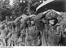 Eight men walking towards the camera with the hands on their heads