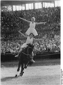 A dark horse, moving in a grassy area. The horse is ridden by two women, one on the others shoulders, and both women are wearing white dresses. In the background is a large seated crowd.