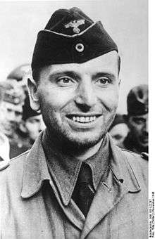 The head and shoulders of a smiling young man. He wears casual army fatigues and is unshaven.