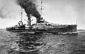 A large gray warship sails through calm seas; a dark cloud of smoke pours out of its two smoke stacks.