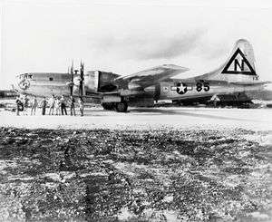 A shiny metal four-engined aircraft stands on a runway. The crew pose in front of it.