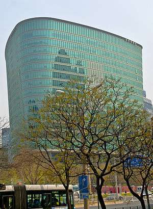 A triangular greenish glass-faced building, flaring outward the higher it gets, rises over a street lined with freshly budding trees on which a blue and white trolleybus is stopped