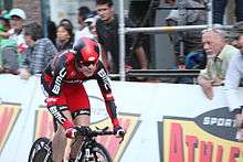 A road racing cyclist in a red, black, and white skinsuit. Spectators watch from behind roadside barricades.