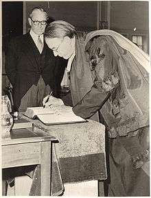 A black-and-white photograph of two men wearing glasses, the man on the right writing in a book while the man on the left watches