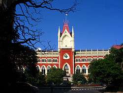 A red-and-yellow building with multiple arches and towers standing against a backdrop of blue sky and framed by trees