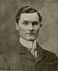 A dark-haired man in his early thirties wearing a high-collared shirt and light-colored jacket, facing left