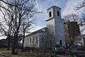 A gray church stands next to an old cemetery. The church has a tower but no steeple, and a circular drive in front.